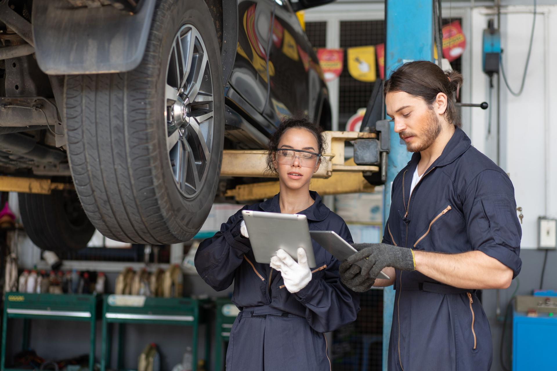 Mechanics man and woman in uniform look at digital tablet discussing car check and repair maintenance in auto service garage