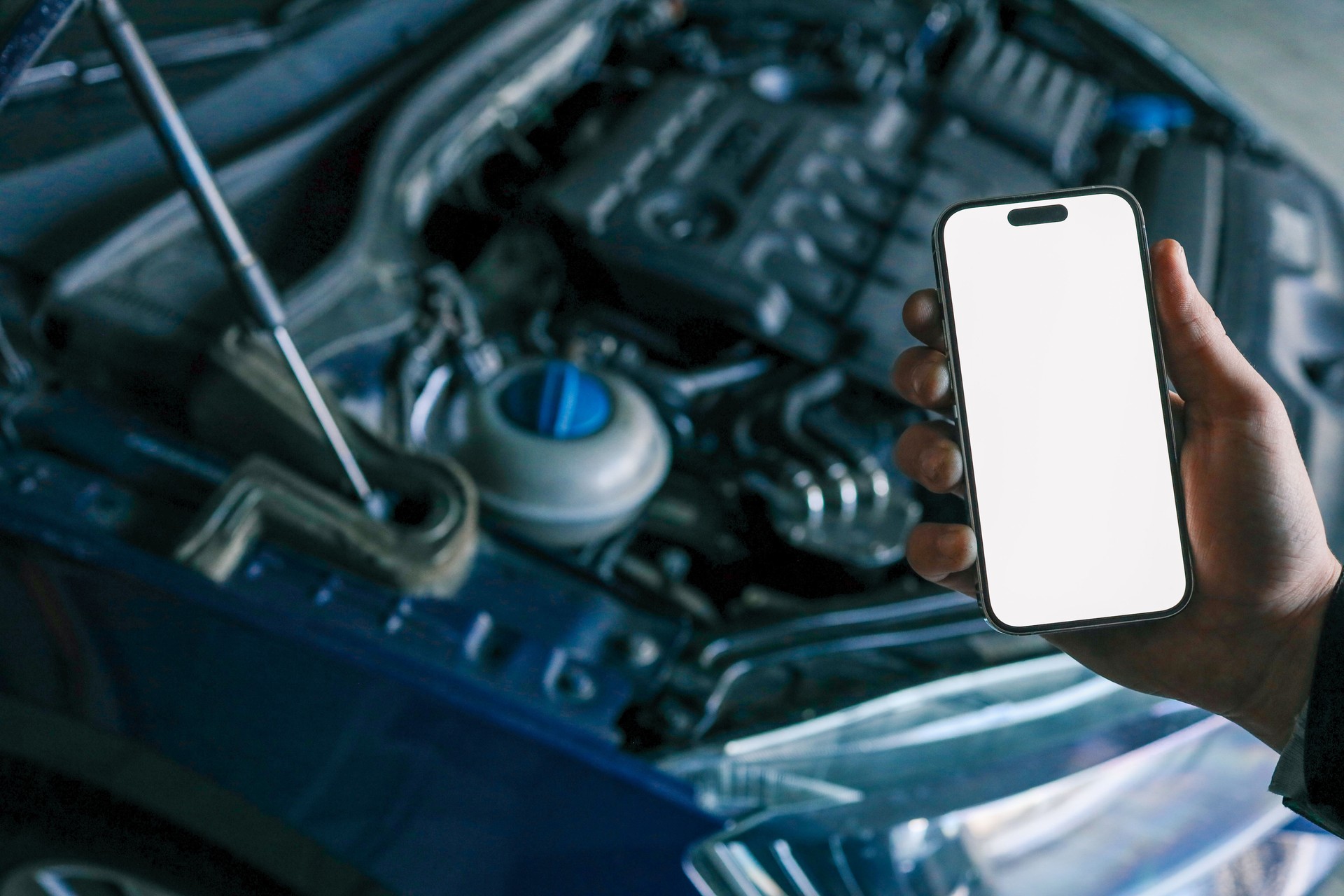 Hand holding a smartphone with a blank screen in front of an open car hood, showing the engine compartment. Concept for car diagnostics, automotive apps, or remote vehicle monitoring
